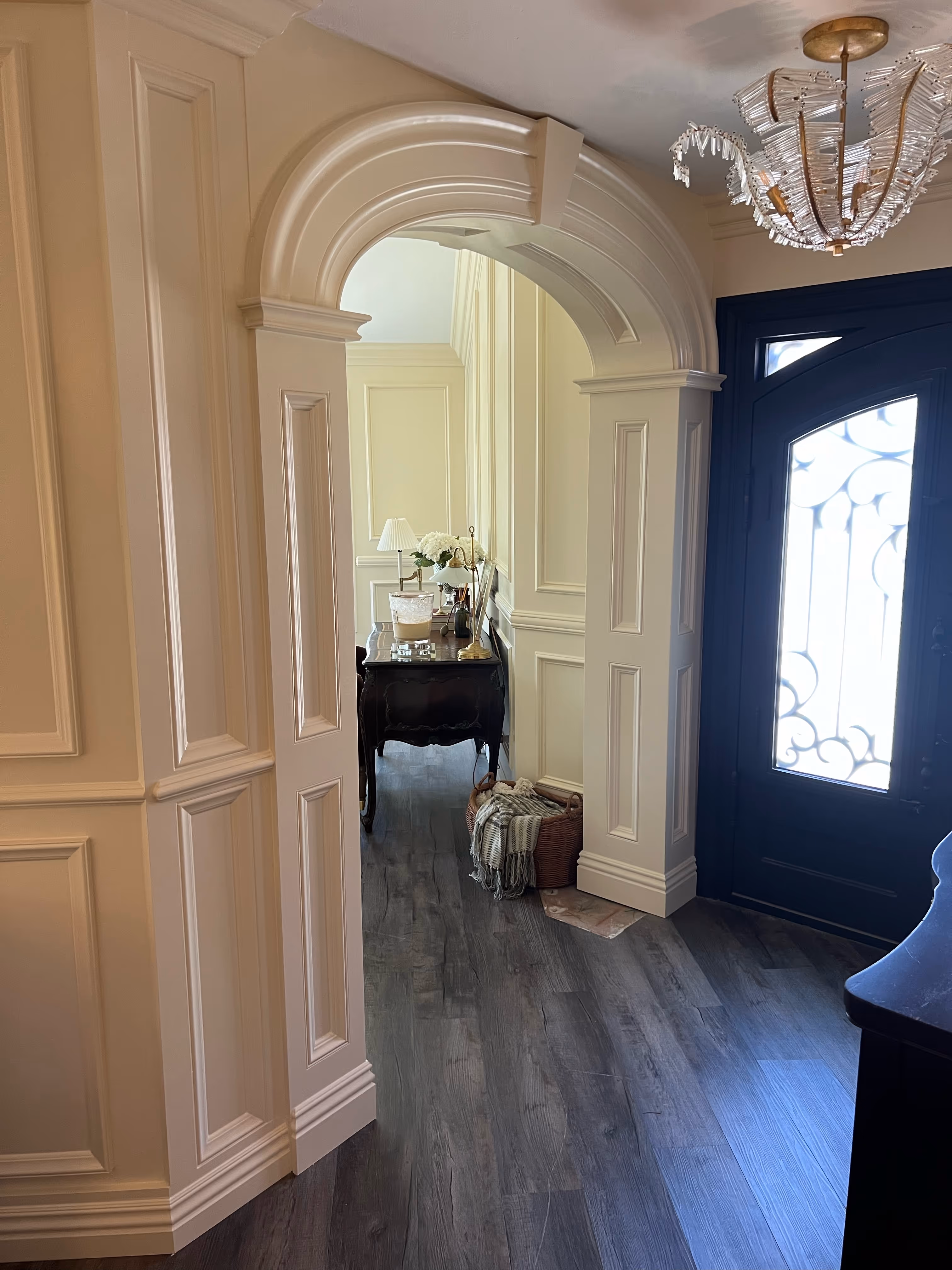 Elegant home entryway with ornate arched doorway, raised panel wainscoting, navy front door with wrought iron glass insert, and crystal chandelier