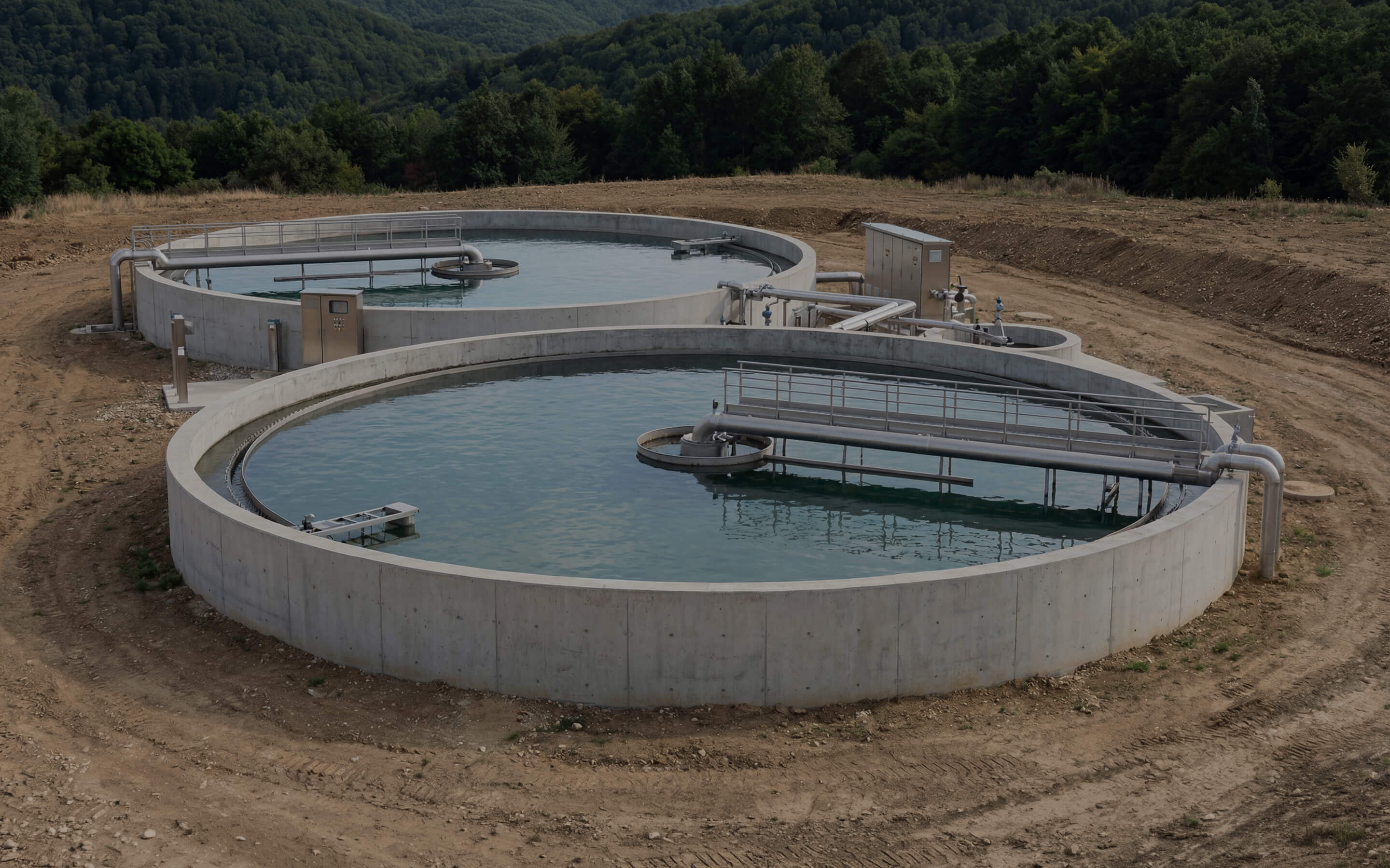 Two large circular concrete water treatment tanks outdoors surrounded by dirt and trees.