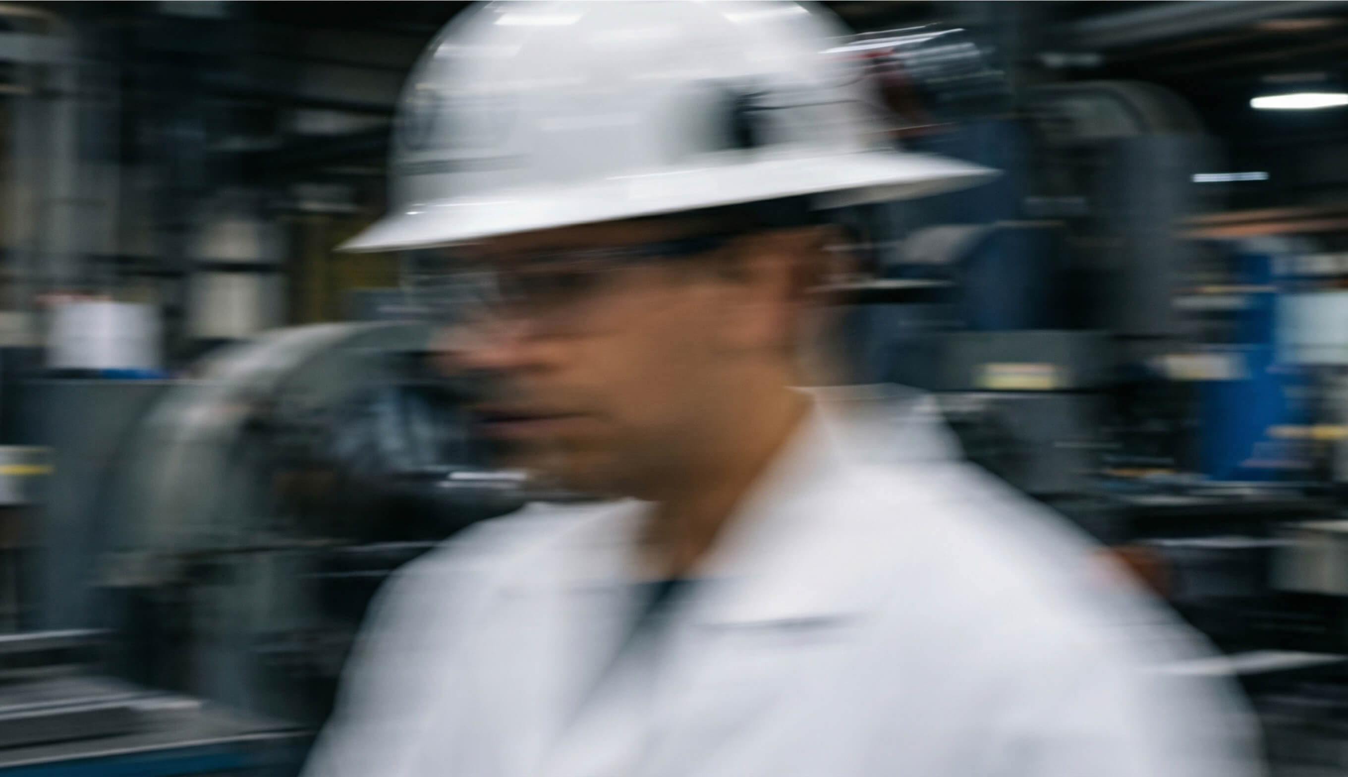 Blurred image of a worker wearing a white hard hat and lab coat in an industrial setting.