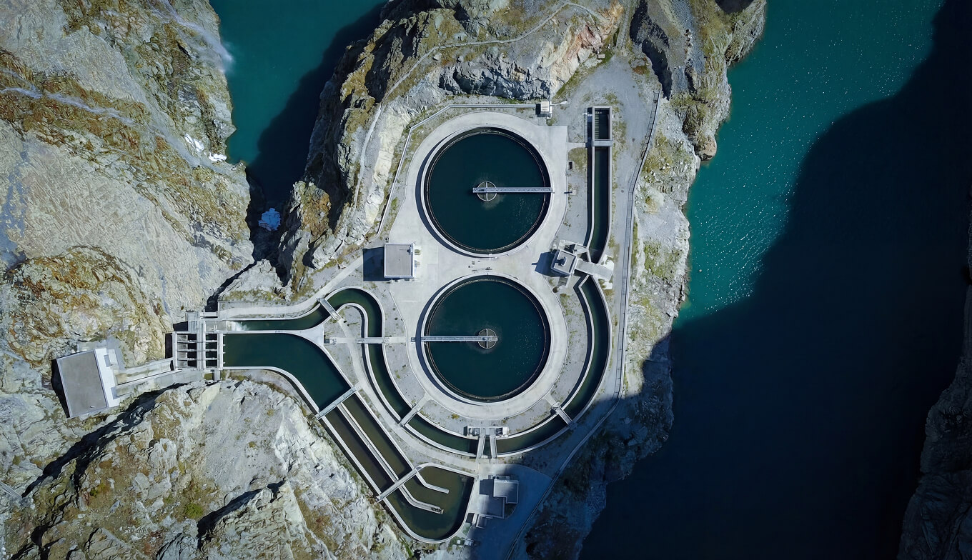 Aerial view of a water treatment facility with circular reservoirs surrounded by rocky terrain and adjacent water body.
