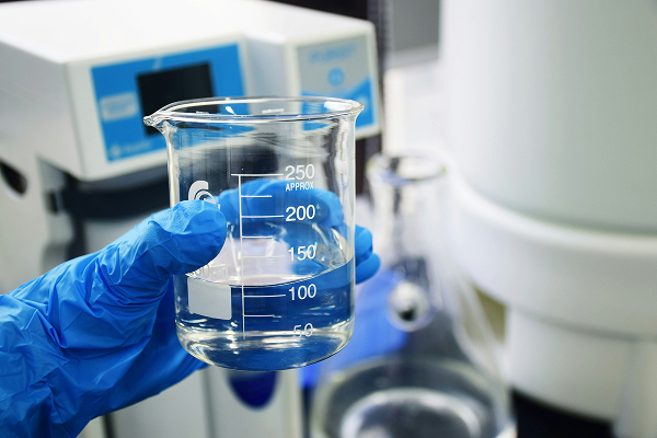 Gloved hand holding a glass beaker filled with clear liquid in a laboratory setting.
