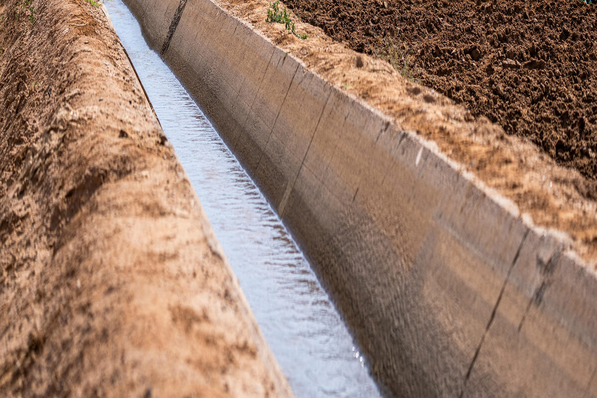 Concrete irrigation canal channeling water beside a cultivated agricultural field.