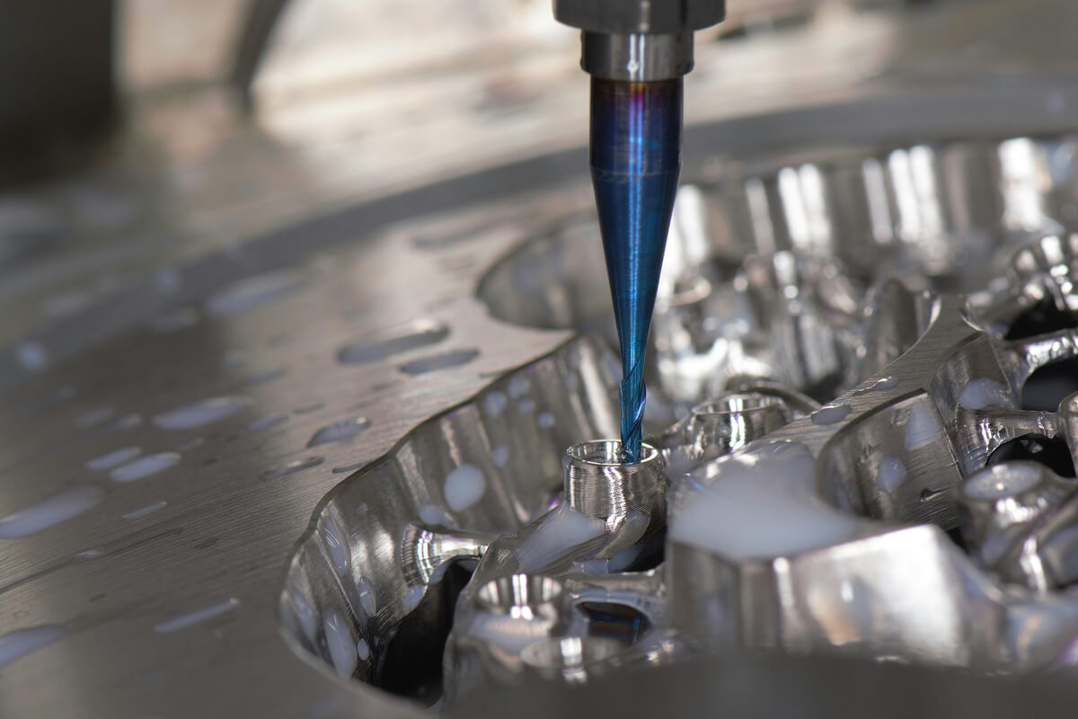 Close-up of a blue drill bit machining a shiny metal part with coolant fluid.
