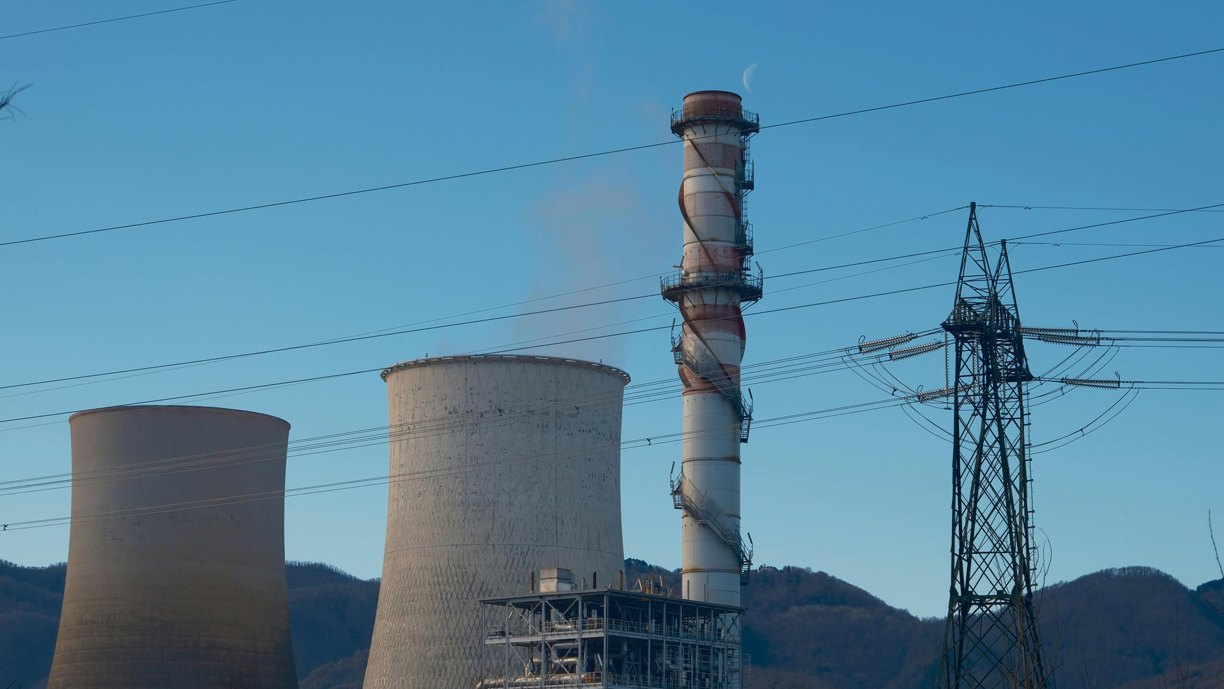 Industrial plant with cooling towers, a tall smokestack, power lines, and mountains in the background.