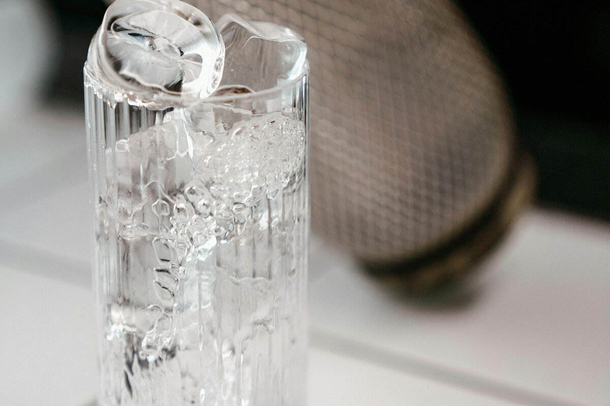 Clear textured glass filled with ice cubes on a white surface with a blurred netted brown object in the background.