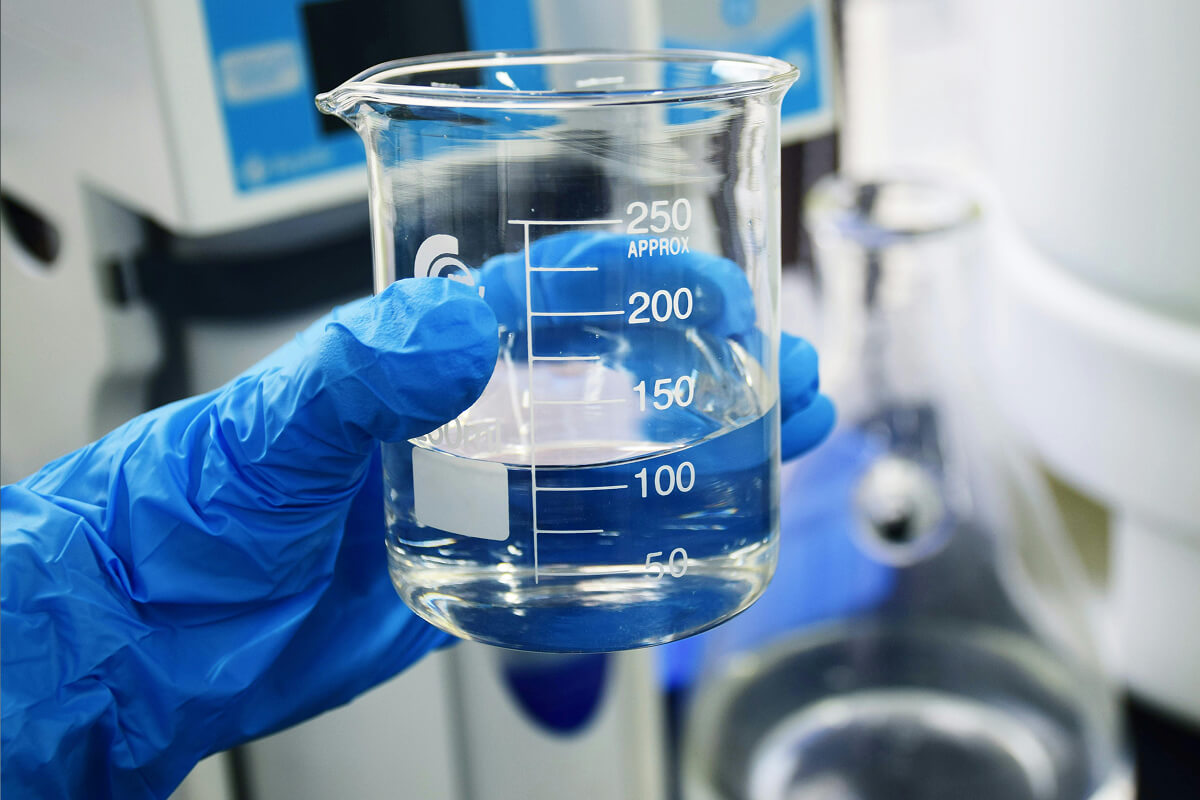 Hand in blue glove holding a glass beaker filled with clear liquid up to about 100 milliliters.