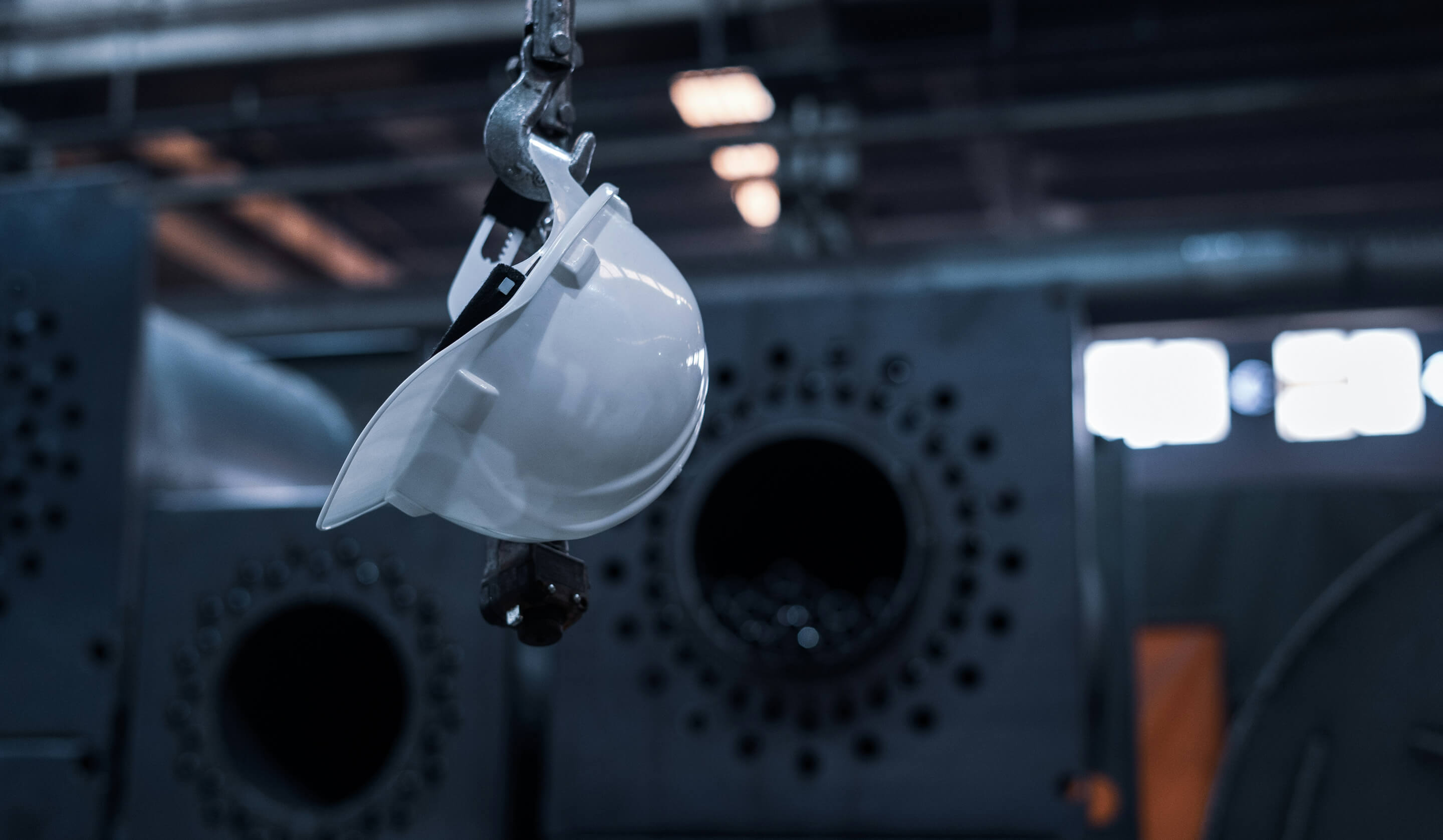 White construction hard hat hanging from a metal hook in a factory setting.