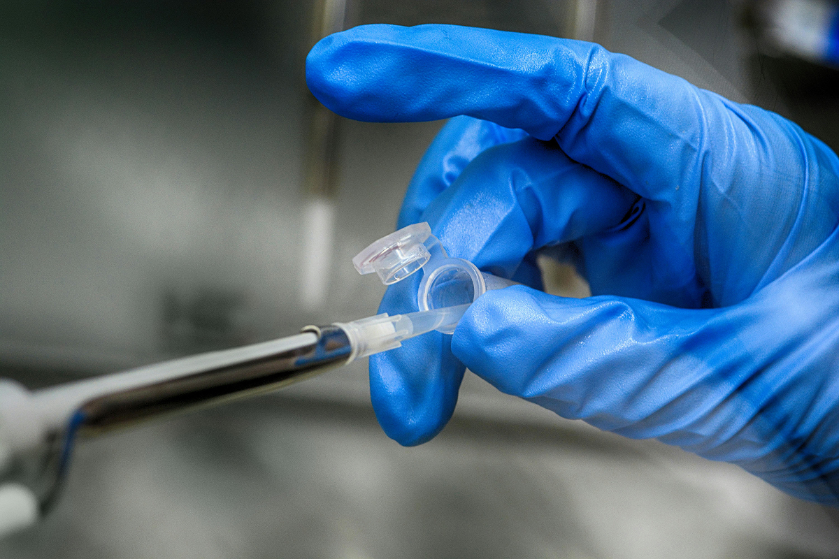 Hand in blue glove using a pipette to transfer liquid into a small plastic vial in a lab setting.