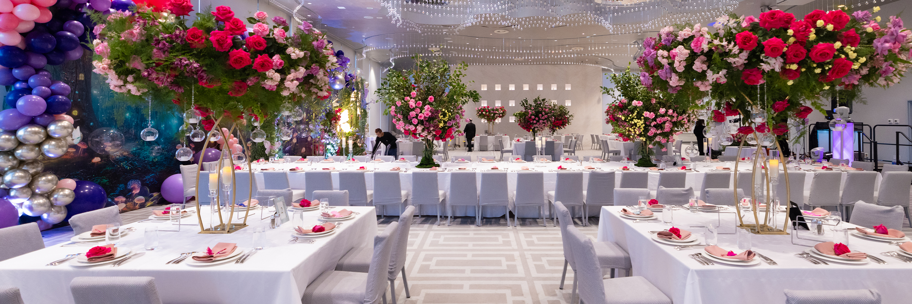 Person sitting cross-legged wearing a colorful jacket and orange pants against a purple background.