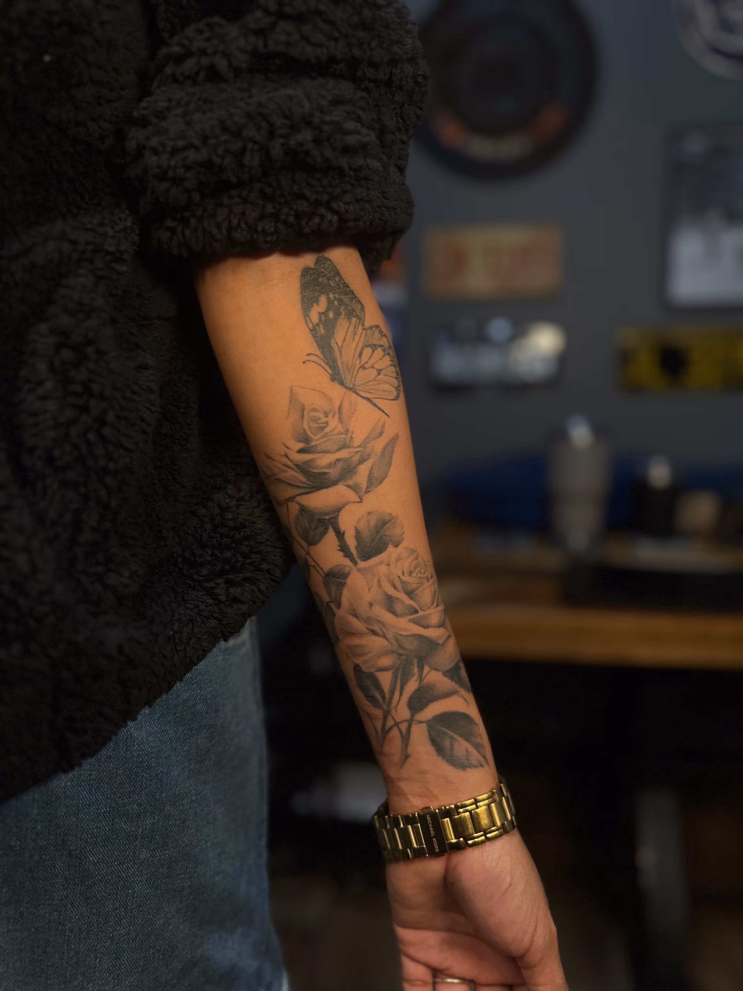 Forearm tattoo of two roses and a butterfly above them on a person wearing a gold watch and dark clothing.