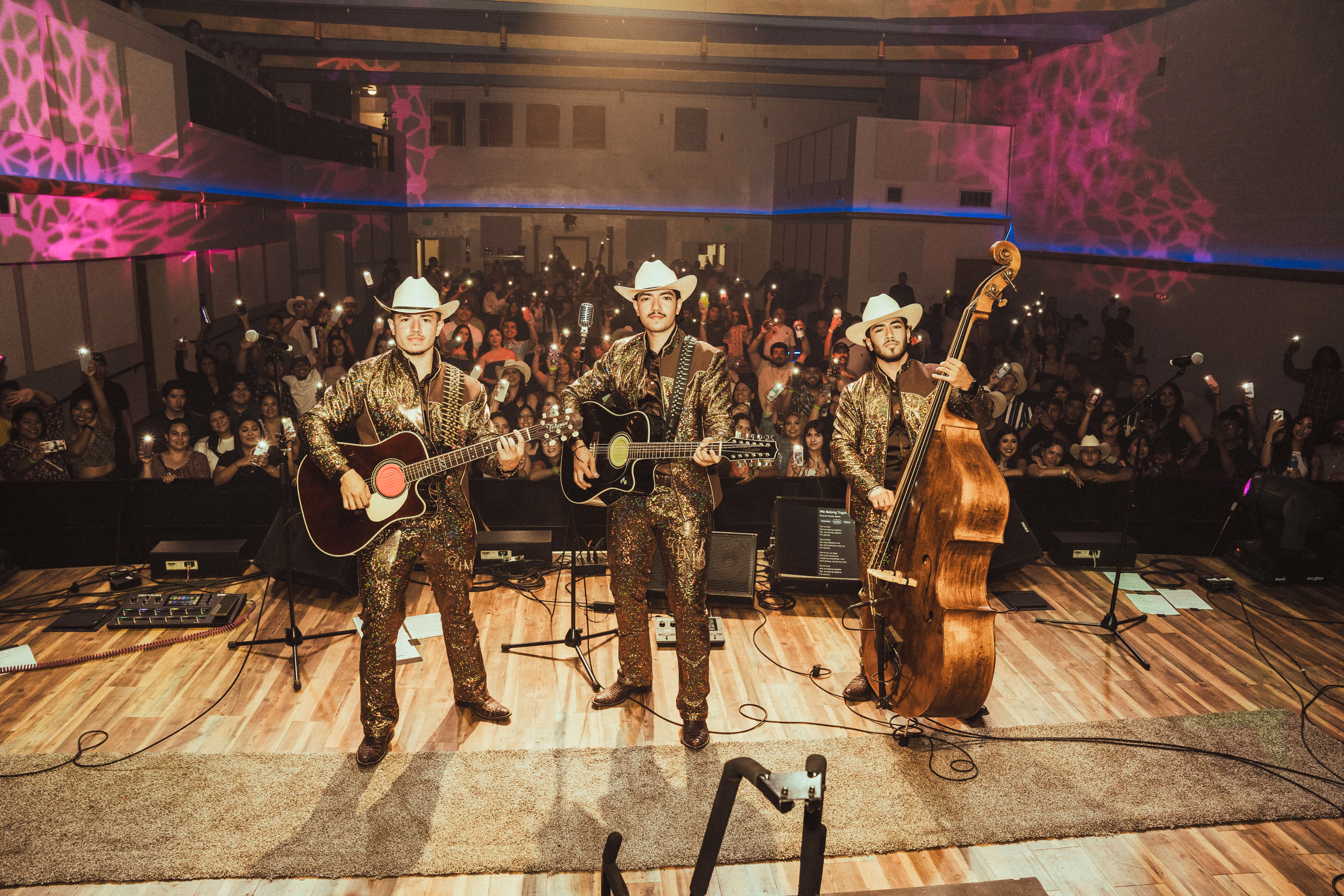 Three musicians in matching sparkling outfits and white cowboy hats play guitars and upright bass on stage in front of an audience with phone lights on.
