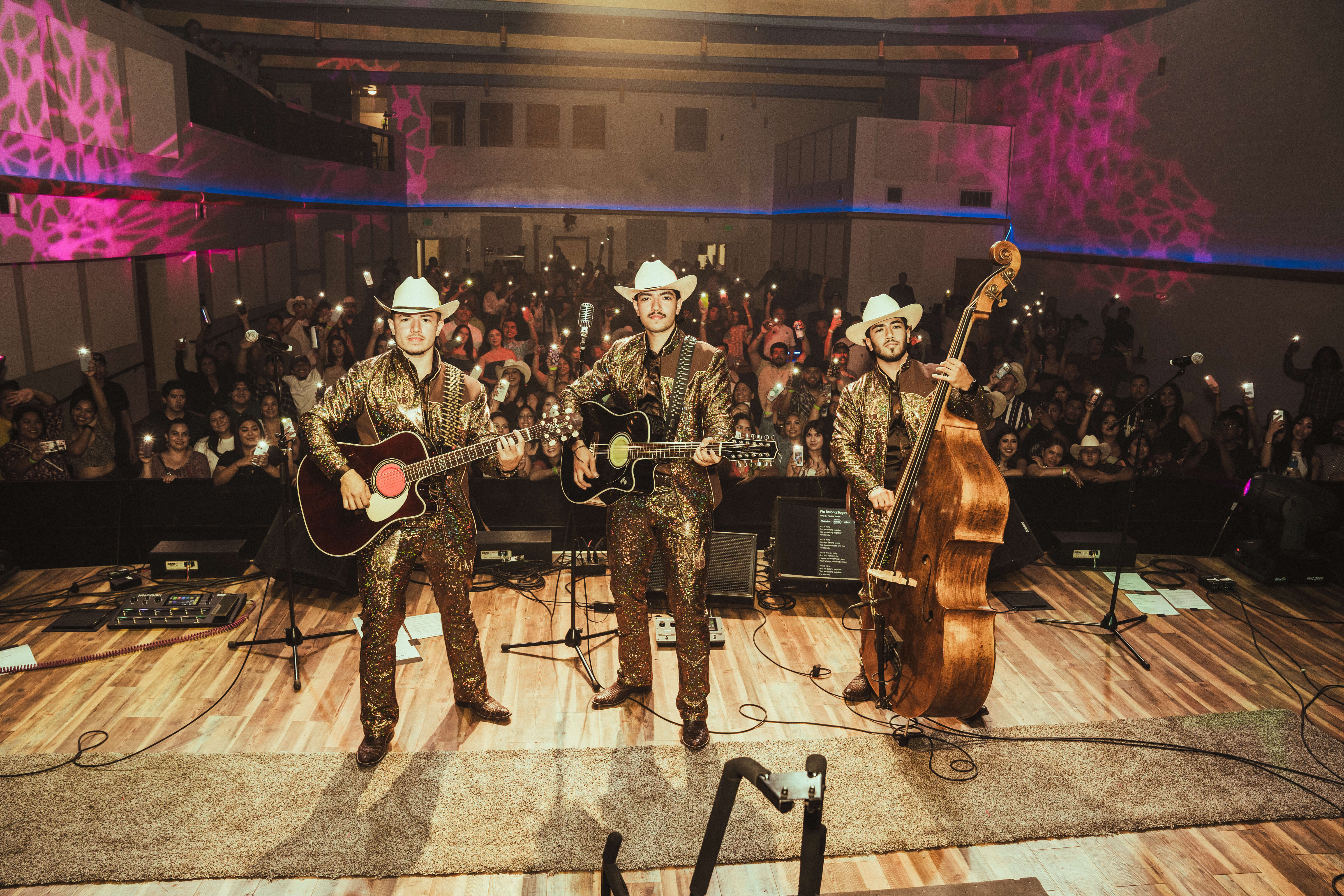 Three musicians in matching sparkling outfits and white cowboy hats play guitars and upright bass on stage in front of an audience with phone lights on.
