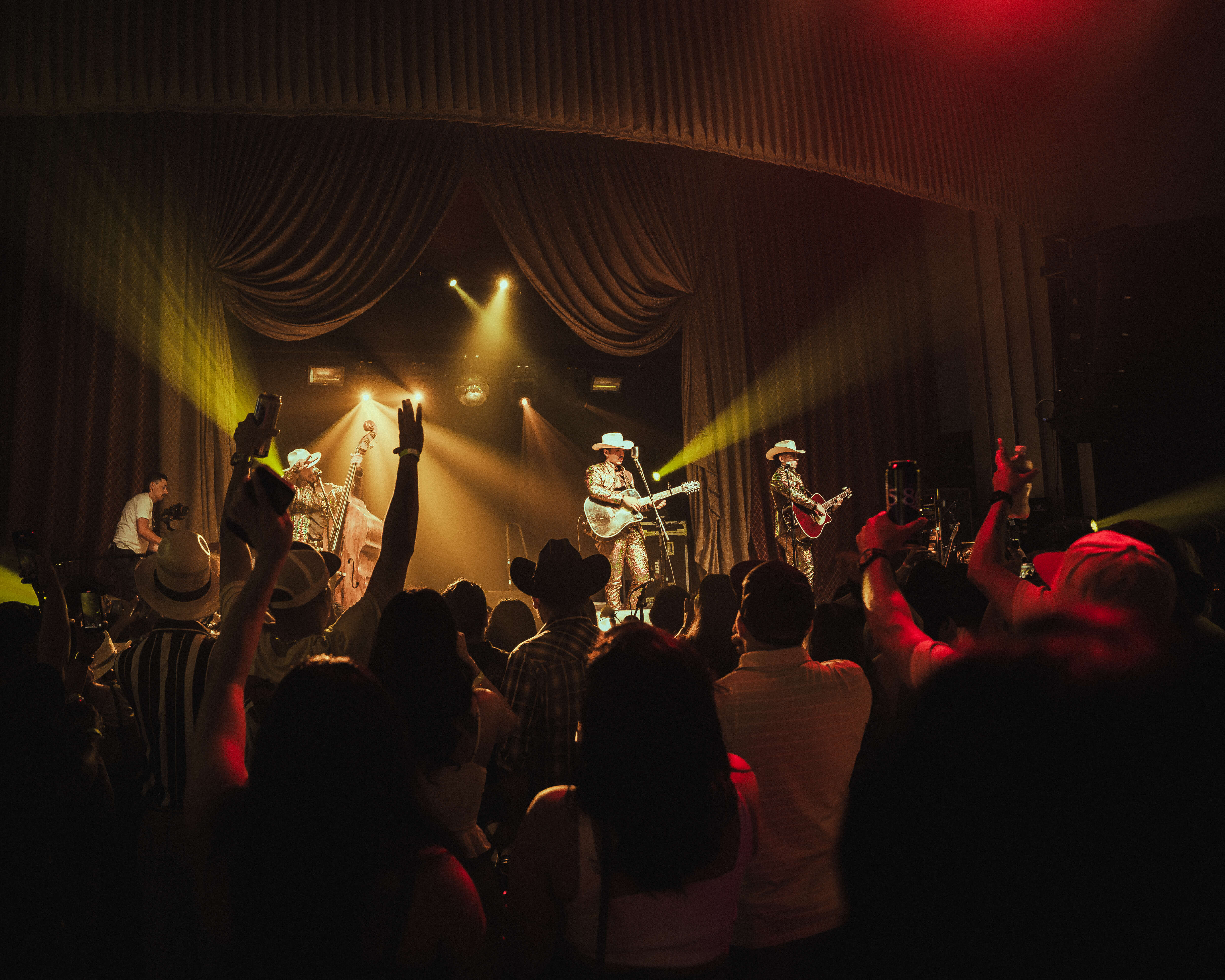 Crowd with raised hands enjoying a live country music concert with three guitarists wearing cowboy hats on stage under yellow spotlights.