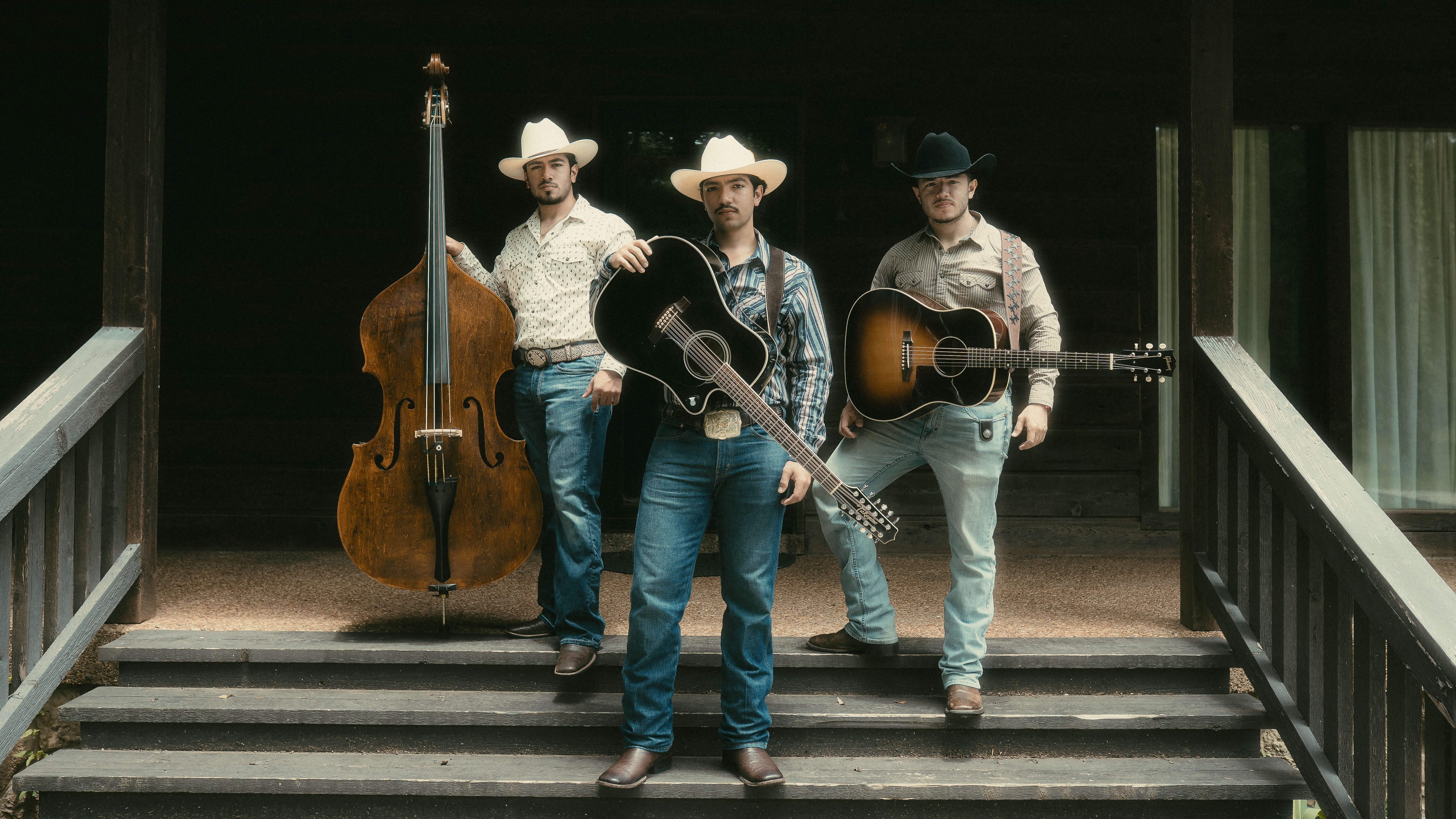 Three men wearing cowboy hats and casual western clothes holding musical instruments including a double bass and two guitars on wooden porch steps.