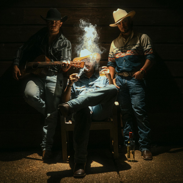 Three men in cowboy hats and western attire, one playing guitar, one sitting smoking a cigar with a drink, and one standing next to a bottle on the ground against a dark wooden wall.