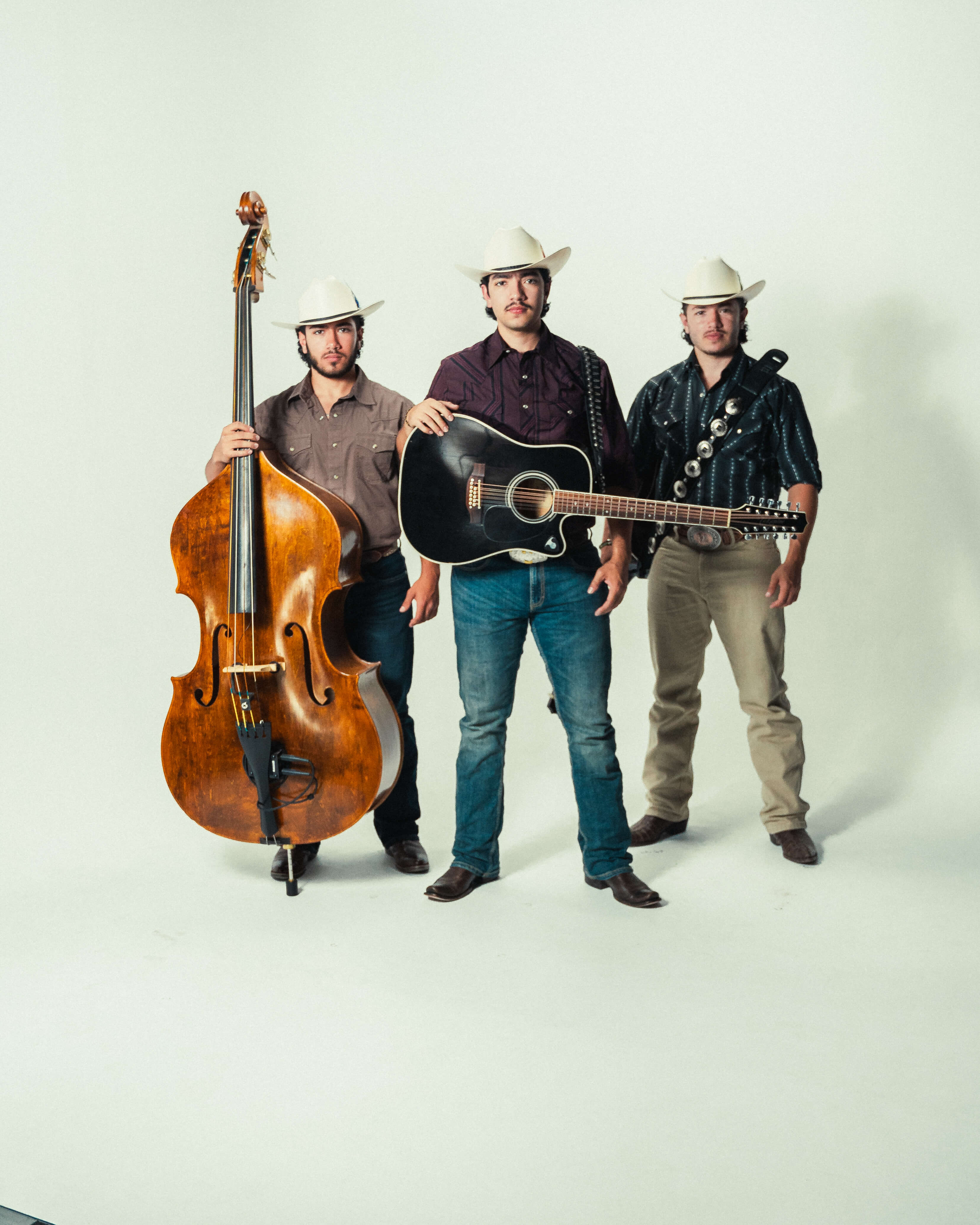 Three men wearing cowboy hats and western shirts posing with a double bass, an acoustic guitar, and a guitarron.