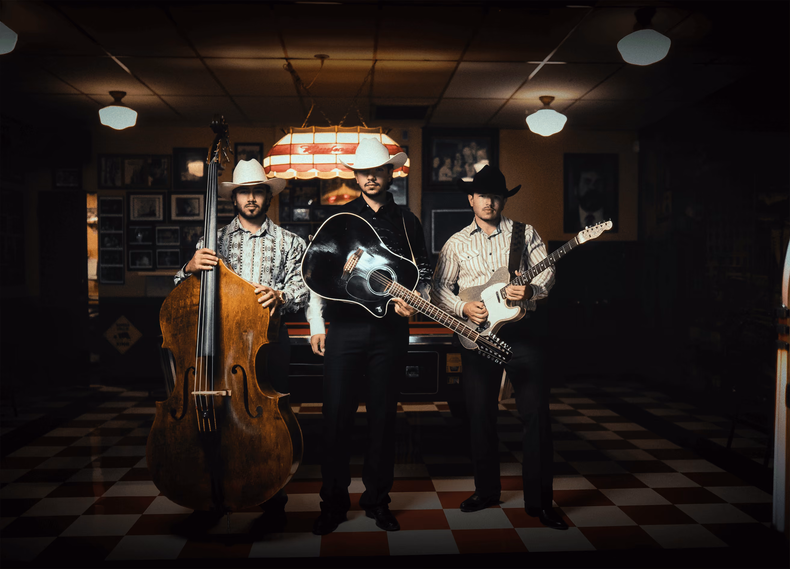 Three men wearing cowboy hats playing string instruments in a dimly lit room with checkered floor.