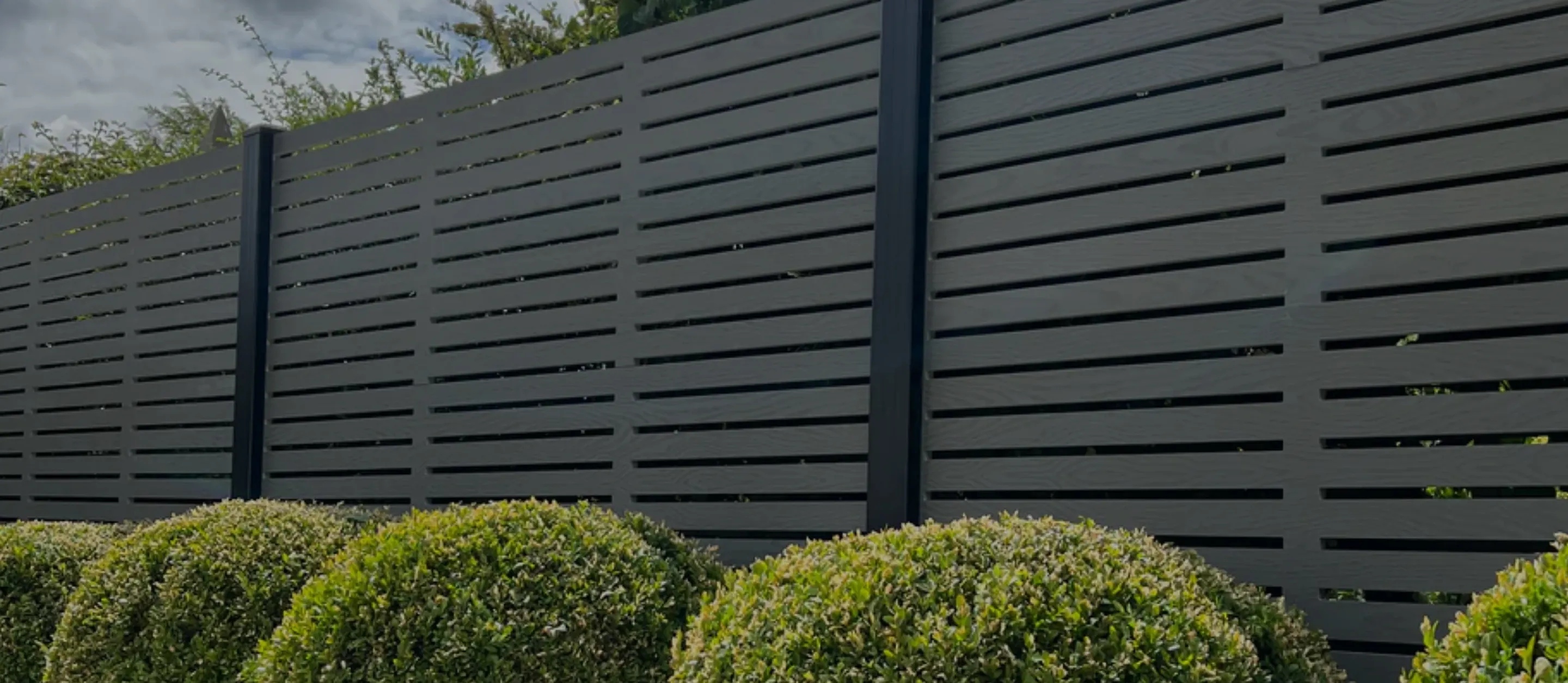 Modern gray horizontal slat fence with green rounded shrubs in front under cloudy sky.