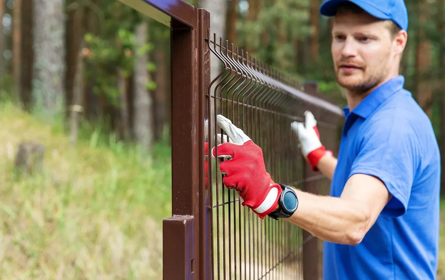 Man wearing red gloves and a blue shirt installing or inspecting a brown metal fence outdoors.