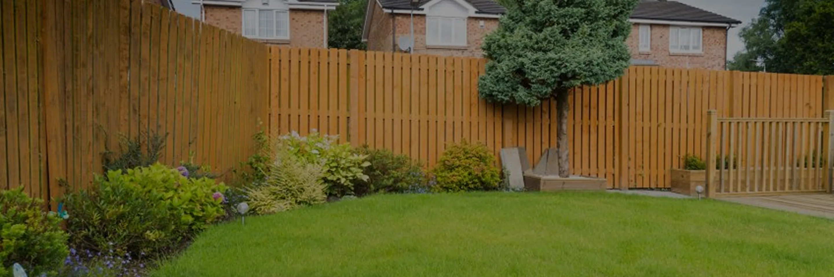 Well-maintained backyard with green grass, a variety of shrubs along a wooden fence, a trimmed tree, and a wooden deck with a gate.