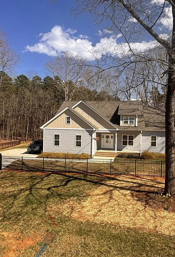Light gray two-story house with a gabled roof, black metal fence in front, and a car parked in the driveway, surrounded by trees under a blue sky with clouds.