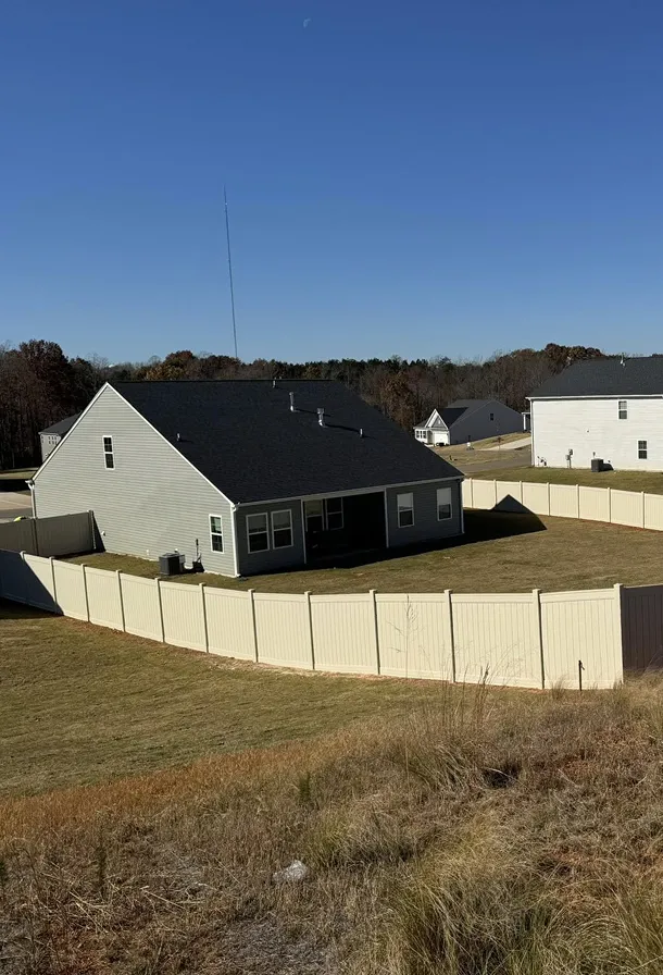Backyard of a gray house with a black roof enclosed by a white fence under a clear blue sky.