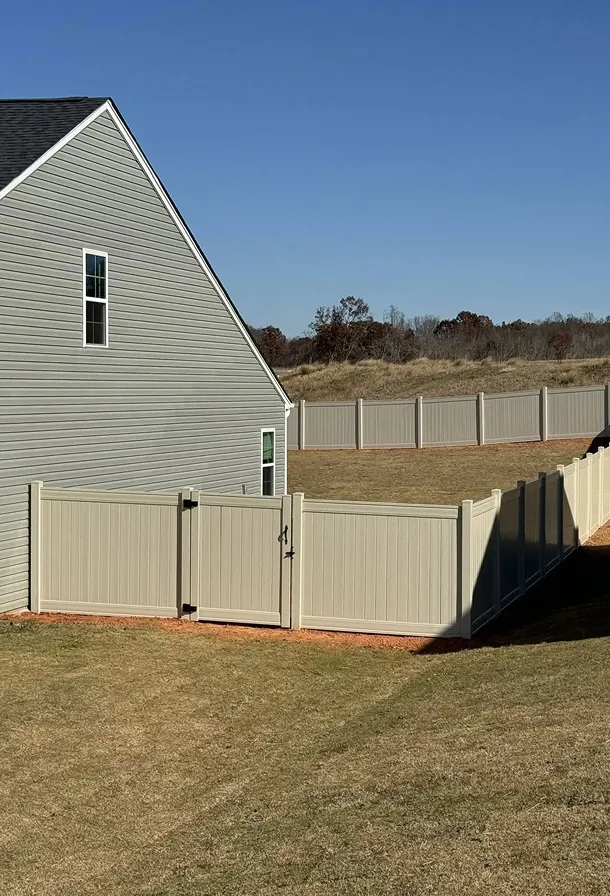Grey house with white trim next to a beige vinyl privacy fence enclosing a yard under clear blue sky.