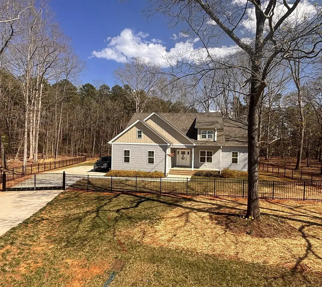 Single-story house with beige siding and a shingled roof, surrounded by a black metal fence and leafless trees under a blue sky with clouds.