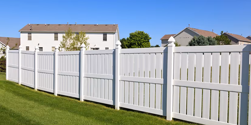 Long white vinyl privacy fence on green lawn with houses and trees in the background under a clear blue sky.
