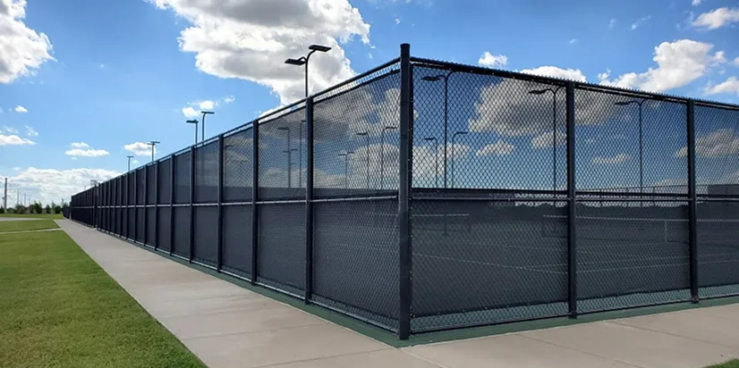 Outdoor tennis courts surrounded by a tall black chain-link fence under a blue sky with scattered clouds.