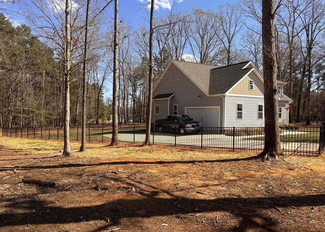 Gray two-story house with attached garage and black pickup truck parked in driveway surrounded by black metal fence and leafless trees.