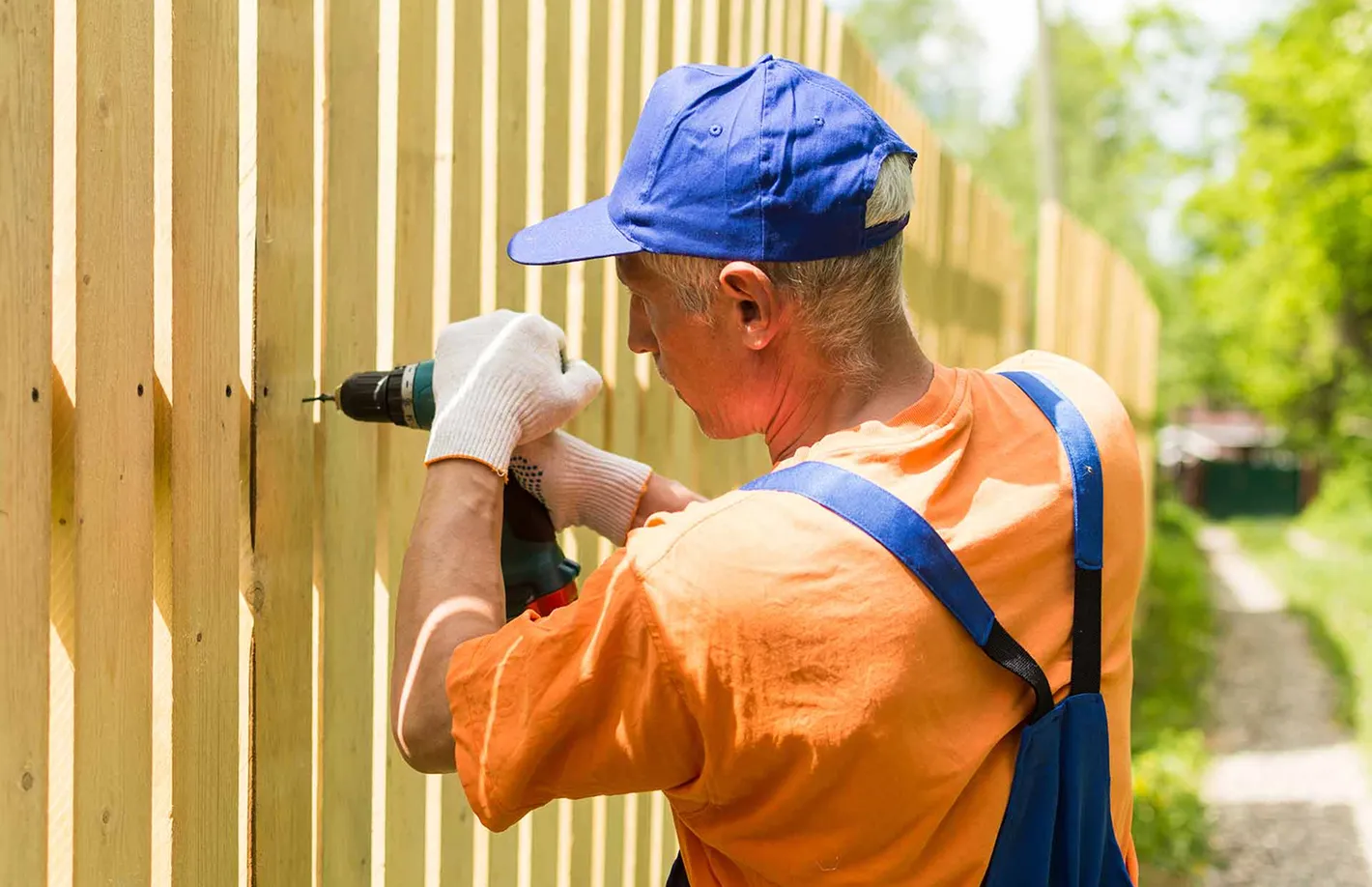 Man in orange shirt and blue cap using a drill to install a wooden fence outdoors on a sunny day.