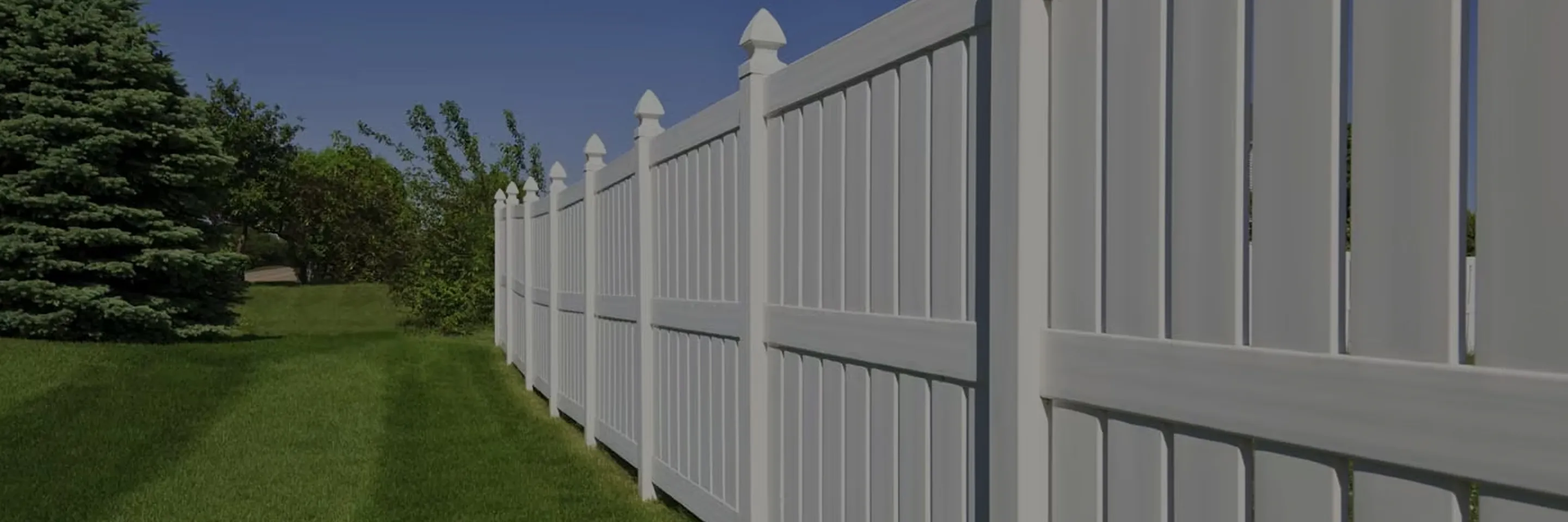Long white vinyl fence with pointed caps along green grassy yard and trees under clear blue sky.