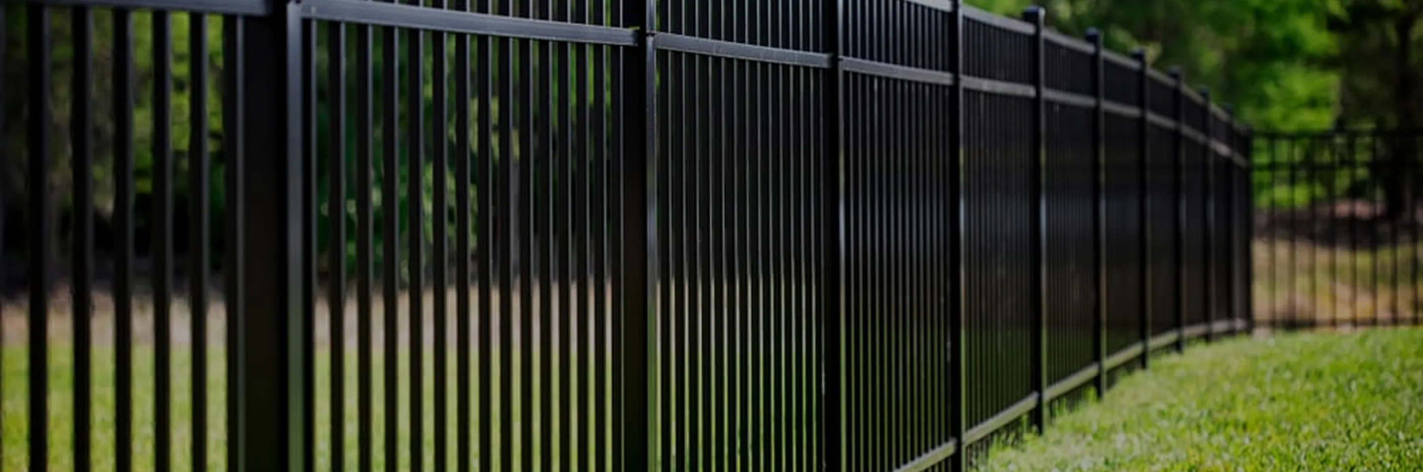 Black metal fence with vertical bars stretching across a grassy area with trees in the background.
