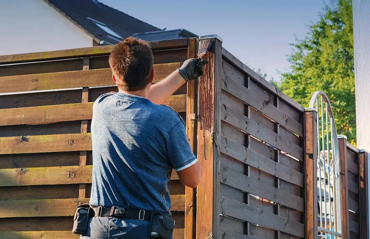 Man wearing gloves and a blue shirt painting a wooden fence outdoors on a sunny day.