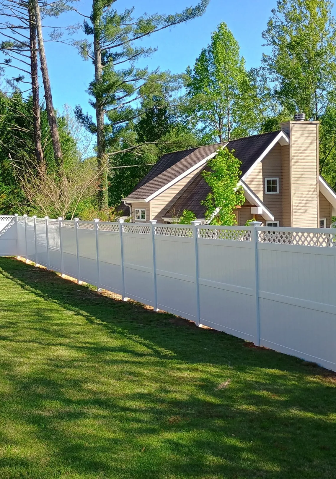 White vinyl fence enclosing a yard with green grass and trees, beige house with a chimney visible in the background.