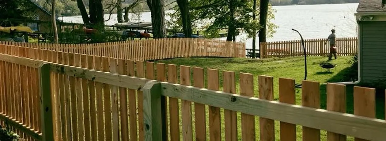 Curved wooden picket fence enclosing a grassy yard with trees and a lake in the background.