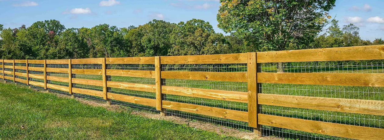 Long wooden fence with horizontal planks and wire mesh on a grassy field with trees in the background under a blue sky.