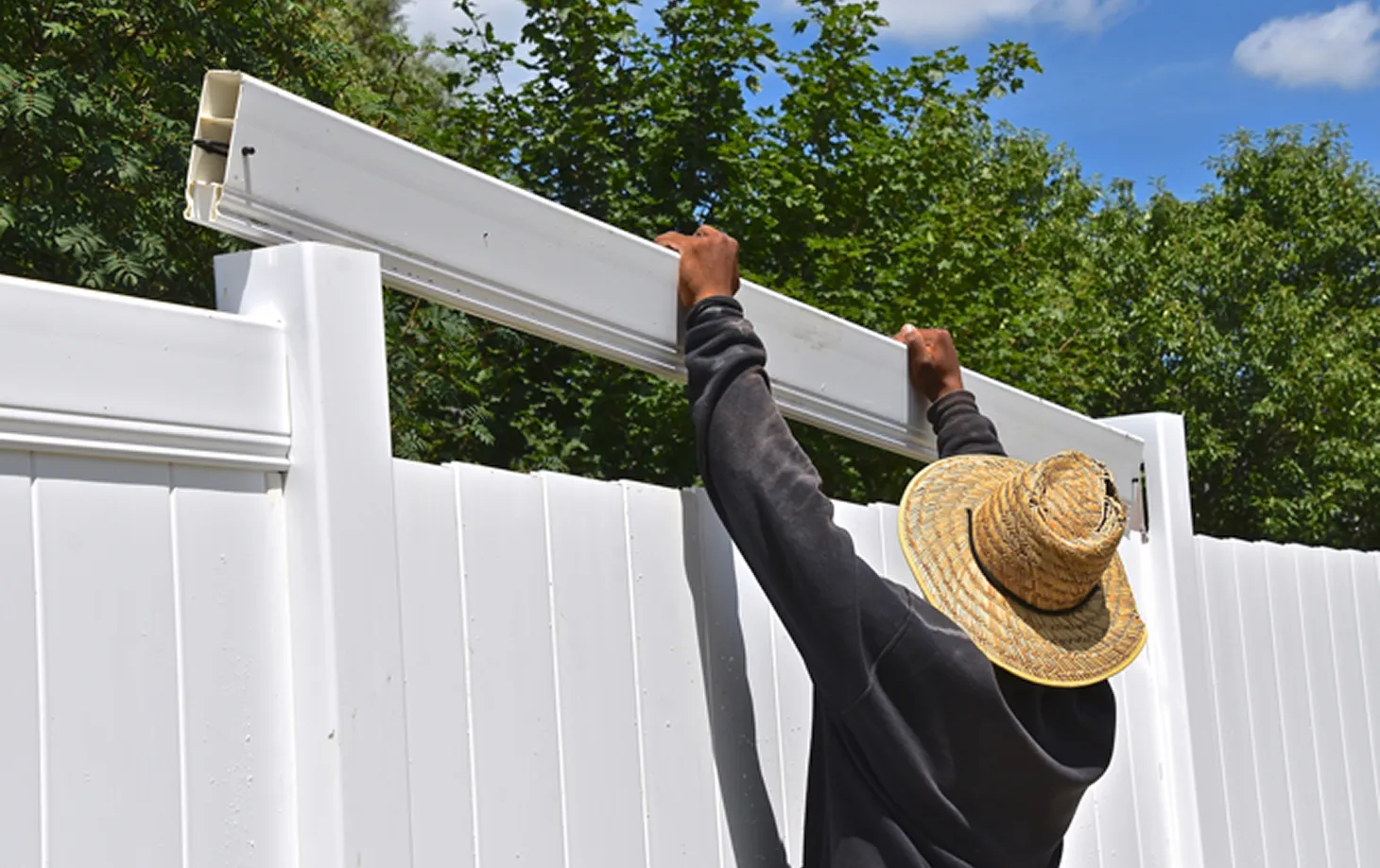 Person wearing a straw hat and dark sweatshirt installing a white vinyl fence panel outdoors with green trees in the background.