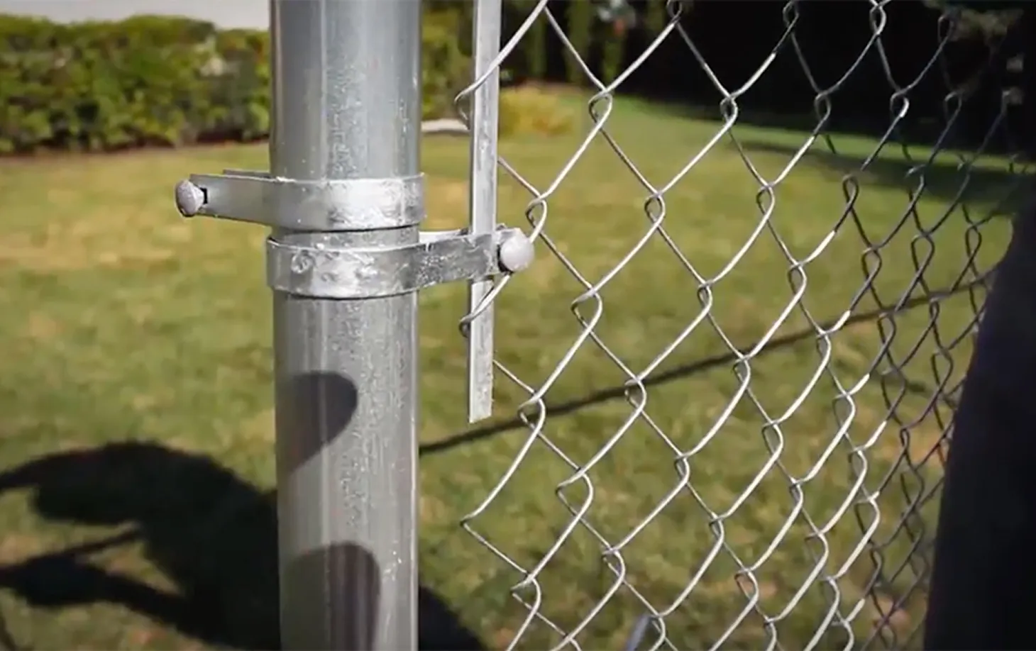 Metal chain-link fence gate latch attached to a vertical post with green grass and bushes in the background.