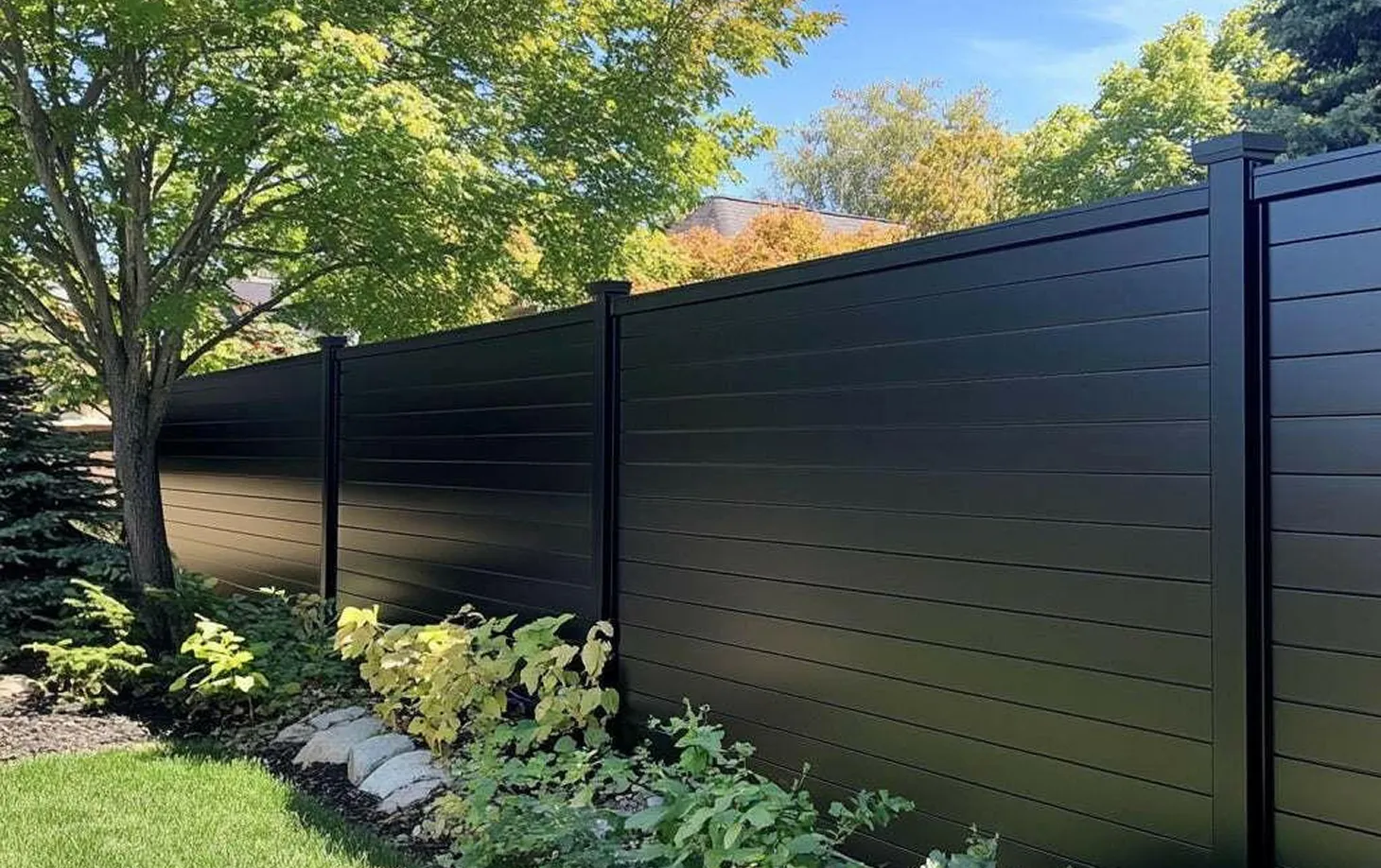 Black aluminum privacy fence panels along a landscaped garden with green trees and plants under a blue sky.