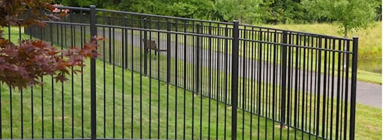 Black metal fence enclosing a grassy area with a paved path and greenery in the background.