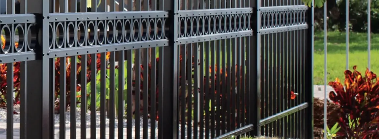 Black metal fence with vertical bars and decorative circular pattern near the top, surrounded by plants and greenery.