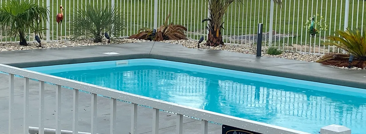 Rectangular blue swimming pool surrounded by a concrete patio and white metal fence with decorative plants outside the fence.