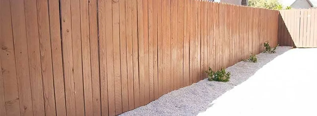 Long wooden fence with a fresh stain on the left side and gravel with small plants along its base.
