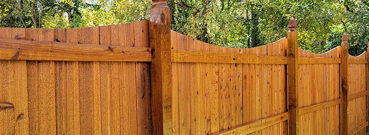 Section of a wooden privacy fence with pointed posts and curved top edges set against green trees.
