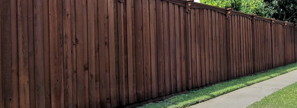 Long, dark brown wooden fence with vertical planks and decorative posts alongside a concrete sidewalk and green grass.