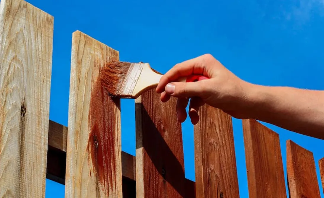 Hand applying brown stain on wooden fence boards with a paintbrush under a clear blue sky.