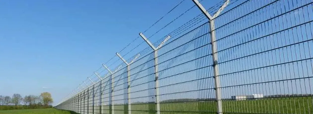 Long tall metal security fence topped with angled barbed wire under a clear blue sky.