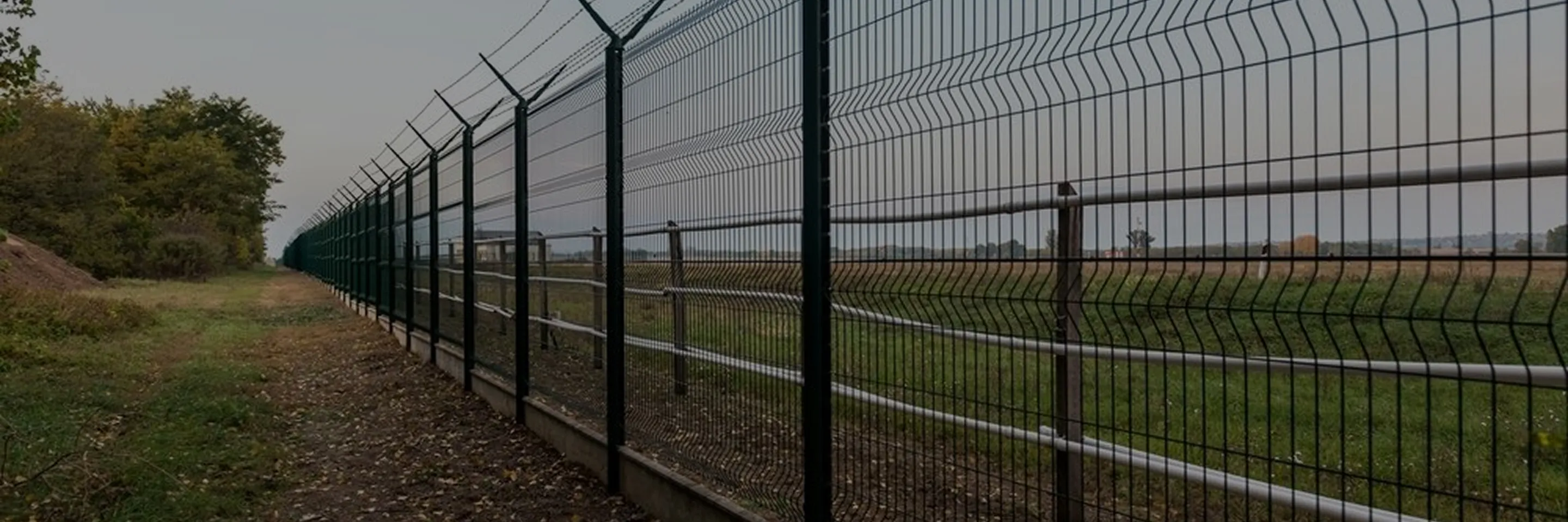 Long black metal security fence with barbed wire on top running along a grassy path and open field.